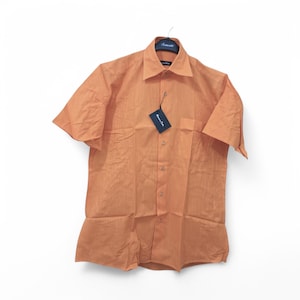 以下が含まれることがあります： 左胸にポケットが付いたオレンジ色の半袖ボタンダウンシャツ。シャツは織り地で、ブランドタグには「Momentum Club」と書いてあります。