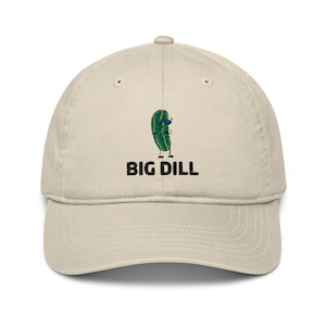 May include: A beige baseball cap featuring the text "BIG DILL" below a cartoon pickle wearing sunglasses. The pickle is green with black legs and arms. The cap is made of a soft material.