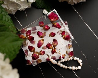 May include: Clear square coasters with dried red rose buds embedded in resin. A white pearl bracelet rests nearby. The coasters are on a black and white marble surface, with white flowers in the background.