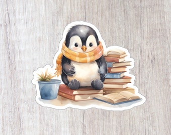 May include: A cute penguin wearing a yellow and orange scarf sits on a stack of books. A small potted plant is to the left of the penguin.