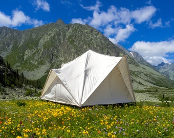 May include: A white canvas tent with a geometric, multi-faceted structure sits in a vibrant meadow filled with yellow and red wildflowers, set against a backdrop of towering, rugged mountain peaks under a bright blue sky with scattered clouds.