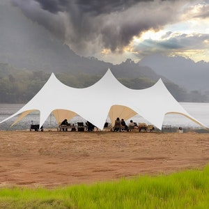 May include: A large white tent with a unique star-shaped design is set up on a sandy area near a lake. Several people are seated under the tent, enjoying the outdoors. The background features a mountain range and a cloudy sky.