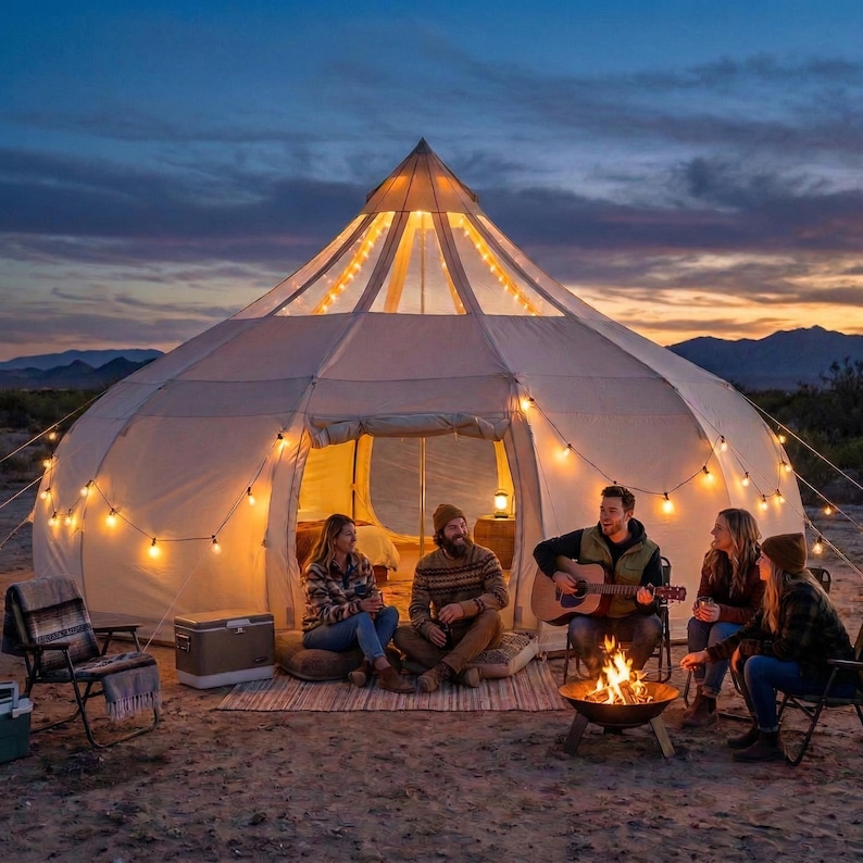 A large white canvas tent with a pointed roof is set up on a sandy beach. A string of lights hangs along the front. People are gathered around, with one playing a guitar. A small table holds food and drinks.