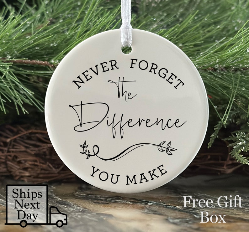 A three-inch flat round ceramic ornament hanging from a white ribbon. There is black text on the front of the ornament that says "Never forget the Difference You Make". There is a decorative flourish separating Difference and You Make.