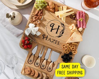 Può includere: Un tagliere per formaggi in bambù personalizzato con un cassetto per coltelli da formaggio. Il tagliere presenta un monogramma "H" per Hazel e include una varietà di formaggi, cracker, uva, olive e pretzel.