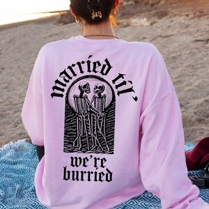 May include: A pink sweatshirt with a black graphic of two skeletons holding hands and the text "married til we're buried".