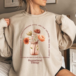 May include: A beige sweatshirt with a floral design and the text "Occupational Therapy" in brown. The text around the floral design reads "Helping you grow to your full potential. Executive functioning, bilateral coordination, social, aids, play, fine motor, visual motor, cognition."