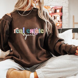 May include: A brown sweatshirt with the text "real estate agent" in colorful letters. The text is in a playful font.