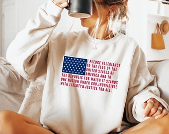 May include: A white sweatshirt with a red, white, and blue American flag graphic and the text "I pledge allegiance to the flag of the United States of America and to the republic for which it stands one nation under God, indivisible with liberty and justice for all."