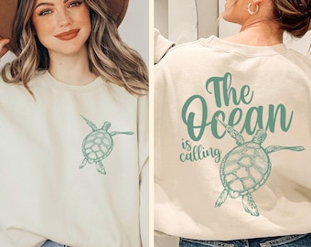 May include: A beige crewneck sweatshirt featuring a small green sea turtle graphic on the front left chest and a large green sea turtle graphic on the back with the text The Ocean is calling in a stylized script font.