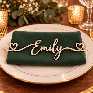 May include: A light-colored wooden name tag featuring the name Emily in a cursive script font, flanked by two small heart shapes on either side, resting on a folded dark green fabric napkin placed on a white plate with a gold rim.