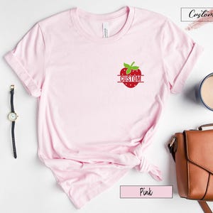 A light pink short-sleeve t-shirt featuring a small graphic of a red strawberry on the left chest area