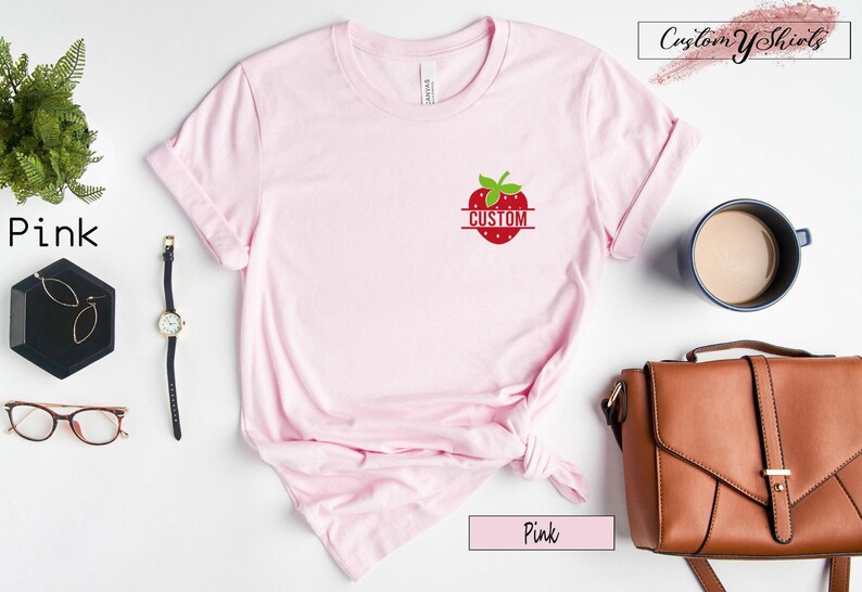 A light pink short-sleeve t-shirt featuring a small graphic of a red strawberry on the left chest area