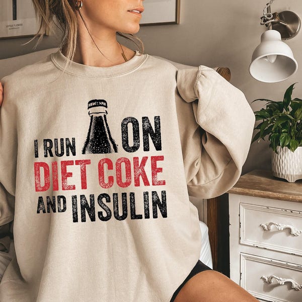 Może przedstawiać: Beżowa bluza z okrągłym dekoltem z nadrukiem graficznym o treści I RUN ON DIET COKE AND INSULIN. Słowa I RUN ON oraz AND INSULIN są czarne, a DIET COKE czerwone. Nadruk zawiera również czarną sylwetkę butelki napoju.