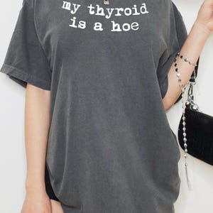 May include: A charcoal gray short-sleeve t-shirt featuring the white text "my thyroid is a hoe" printed in a typewriter-style font across the chest. The garment has a crew neckline and a relaxed, loose-fitting silhouette with a faded, worn-in appearance.