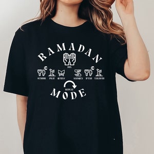 Pu&ograve; includere: Maglietta nera con un design grafico bianco che dice "Ramadan Mode" e include icone che rappresentano le diverse parti della routine quotidiana del Ramadan. Le icone includono una luna crescente, una mano in preghiera, un libro, un piatto con cibo, un orologio e una moschea.