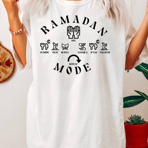 Pu&ograve; includere: Maglietta bianca con testo nero che recita "Ramadan Mode" e include un cerchio di icone che rappresentano diversi aspetti del Ramadan, tra cui suhur, preghiera, Corano, sadaqa, iftar e taraweeh.