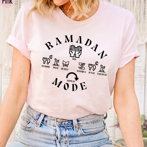 Pu&ograve; includere: Maglietta rosa con il testo "RAMADAN MODE" in nero. Il testo &egrave; circondato da un cerchio di icone che rappresentano le pratiche quotidiane del Ramadan, tra cui suhur, preghiera, Corano, sadaqa, iftar e tarawih.
