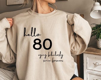 May include: A beige crewneck sweatshirt featuring black text that reads hello 80 and aging fabulously since forever. The garment has long sleeves and a ribbed neckline, constructed from a solid-colored fabric with a clean, simple design.