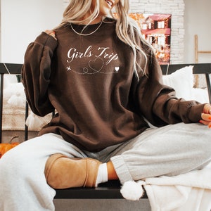 K&ouml;nnte beinhalten: Brauner Pullover mit dem Text "Girls Trip" in wei&szlig;er Kursivschrift, einem kleinen Flugzeug und einem Herzen, die durch eine gepunktete Linie verbunden sind. Der Pullover wird von einer Person getragen, die auf einer Bank sitzt.