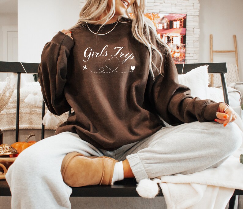 K&ouml;nnte beinhalten: Brauner Pullover mit dem Text "Girls Trip" in wei&szlig;er Kursivschrift, einem kleinen Flugzeug und einem Herzen, die durch eine gepunktete Linie verbunden sind. Der Pullover wird von einer Person getragen, die auf einer Bank sitzt.