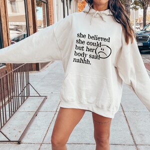 May include: A white hoodie with the text "she believed she could but her body said nahhh." and a black sad face graphic.