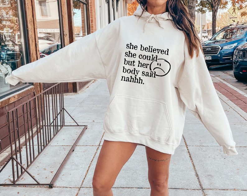 May include: A white hoodie with the text "she believed she could but her body said nahhh." and a black sad face graphic.