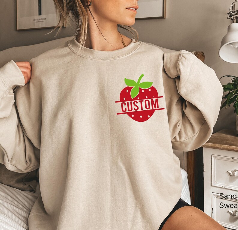 A graphic on the upper left chest, shown on a sand-colored crewneck sweatshirt