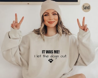 May include: A sand-coloured crewneck sweatshirt featuring the black text IT WAS ME! I let the dogs out. with a paw print icon containing a heart shape. The garment is paired with a matching sand-coloured knit beanie hat, creating a coordinated set.