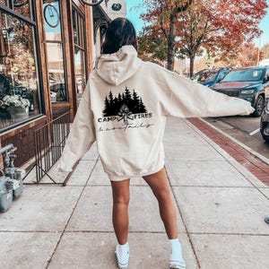 A beige hooded sweatshirt featuring a black graphic on the back. The design displays a silhouette of pine trees above a campfire with the text CAMPFIRES cocktails printed in a combination of bold block and cursive lettering