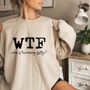 May include: A beige sweatshirt with black text that reads "WTF who's turning fifty?"