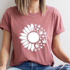 May include: A heather mauve t-shirt with a white graphic of a sunflower with marijuana leaves replacing half of the petals.