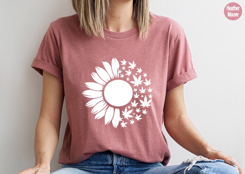 May include: A heather mauve t-shirt with a white graphic of a sunflower with marijuana leaves replacing half of the petals.