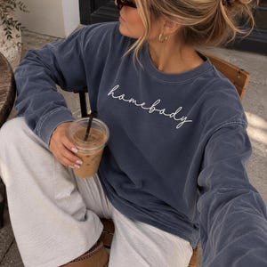 May include: A long-sleeve crewneck sweatshirt in a faded navy blue wash featuring the word homebody printed in white cursive script across the chest. The garment has a relaxed fit with ribbed cuffs and a classic round neckline.