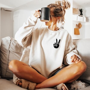 May include: A beige crewneck sweatshirt featuring a black peace sign graphic on the left chest. The long-sleeved pullover has a relaxed fit with ribbed cuffs and a ribbed neckline, providing a simple and clean design for casual daily wear.