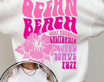 May include: A white long-sleeve sweatshirt featuring a large pink graphic on the back with the text Ocean Beach, Surf Memories, California, Wild Wave, 1971 and a hibiscus flower. A smaller matching pink hibiscus flower is printed on the front.