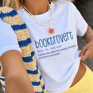May include: A white short-sleeve t-shirt featuring the dark blue text booktrovert, the phonetic pronunciation, and the definition: A person who prefers the company of fictional characters to real people. The shirt is paired with a straw bag with blue stripes.