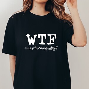 May include: Black oversized t-shirt with white text that reads "WTF who's turning fifty?"