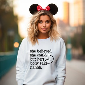 May include: White sweatshirt with black text that reads "she believed she could but her body said nahhh."  A black and white sad face is next to the text. The model is wearing red and white Minnie Mouse ears.