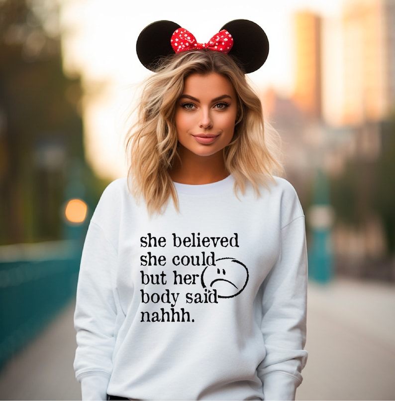 May include: White sweatshirt with black text that reads "she believed she could but her body said nahhh."  A black and white sad face is next to the text. The model is wearing red and white Minnie Mouse ears.