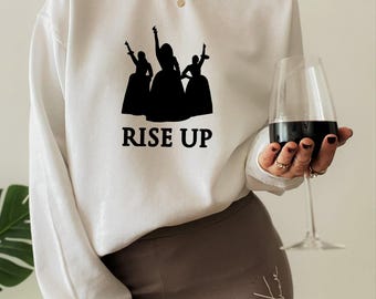 以下が含まれることがあります： 腕を上げたドレス姿の3人の人物の黒いグラフィックと、その下に「RISE UP」という文字がプリントされた白い長袖スウェットシャツ。クルーネックで、袖口はリブ編みになっています。清潔感のある白地です。