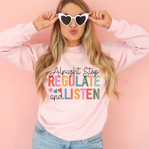 May include: A pink sweatshirt with the text "Alright Stop Regulate and Listen" in colorful letters. The text is in a retro style font with hearts on either side of the word "Regulate".