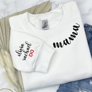 Może przedstawiać: Biała bluza z napisem "mama" w czarnym, ozdobnym pismem na przodzie i napisem "olivia michael" z czerwonym symbolem nieskończoności na rękawie.