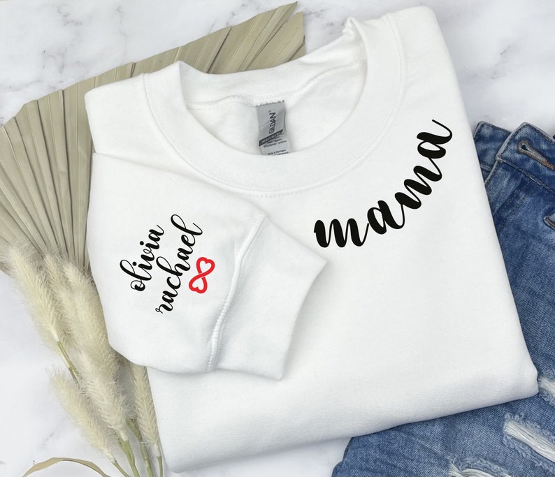 Może przedstawiać: Biała bluza z napisem "mama" w czarnym, ozdobnym pismem na przodzie i napisem "olivia michael" z czerwonym symbolem nieskończoności na rękawie.