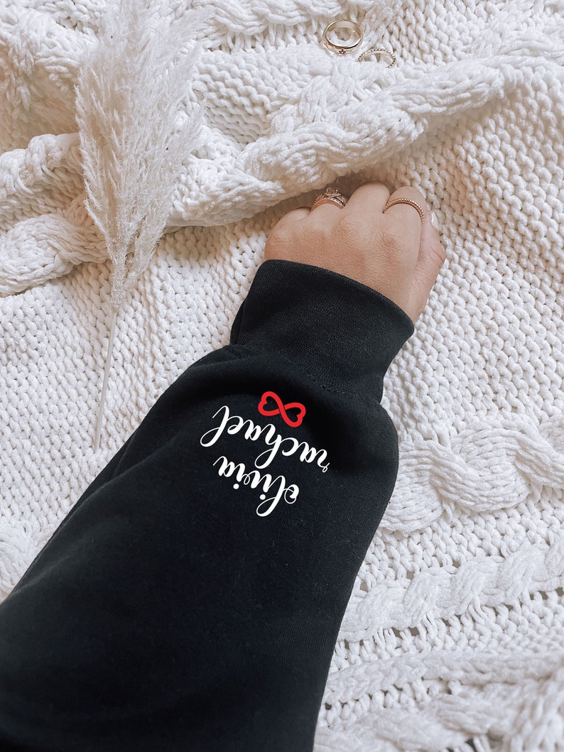 Może przedstawiać: Czarna bluza z białym napisem "Live your magic" z symbolem nieskończoności i czerwonym sercem.