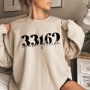 May include: A sand-colored crewneck sweatshirt featuring the black text 33162 and the script text Miami, Florida printed across the chest. The garment has long sleeves, a ribbed neckline, and a relaxed fit, showcasing a simple and clean design.