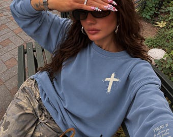 May include: A dusty blue crewneck sweatshirt featuring a white distressed cross graphic on the left chest and the text "Psalms 91: He will cover you with his feathers and under his wings you will find refuge" printed on the left sleeve cuff.