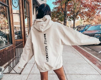 May include: An oversized cream-colored hooded sweatshirt featuring the text "barely tolerable" printed vertically in black lettering down the center of the back. The garment includes a hood and long sleeves, with a relaxed fit and a plain front.