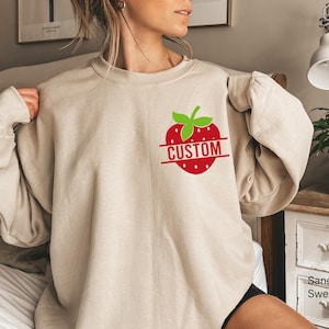 A sand-colored crewneck sweatshirt featuring a graphic on the upper left chest. The graphic displays a red strawberry with green leaves and a horizontal red line across the center containing the white text CUSTOM