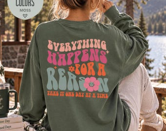 Könnte beinhalten: Ein moosgrünes Langarmshirt mit einem Rückendruck, der den Text Everything Happens For A Reason und Take It One Day At A Time in wellenförmiger, mehrfarbiger Schrift zeigt, akzentuiert durch kleine Sterne und eine rosa Smiley-Blume.